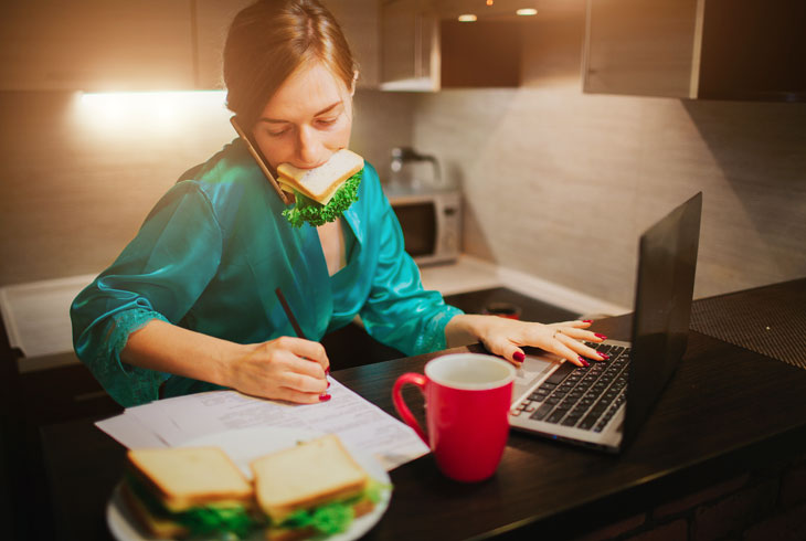Девушка с бутербродом во рту работает за ноутбуком