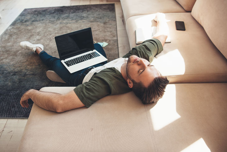 Уставший мужчина лежит на полу с ноутбуком и отдыхает после удалённой работы раскинув руки на диване